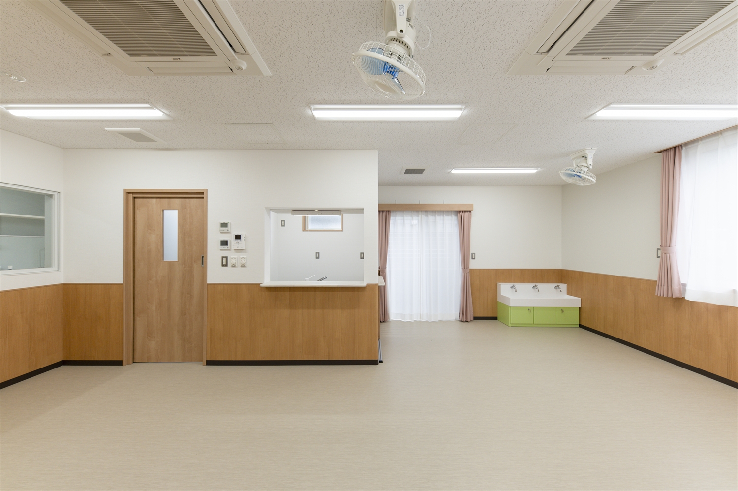 院内保育園の室内写真。木目調の腰壁と扉、明るい窓、子ども用手洗いが配置された清潔で安心感のある保育室。