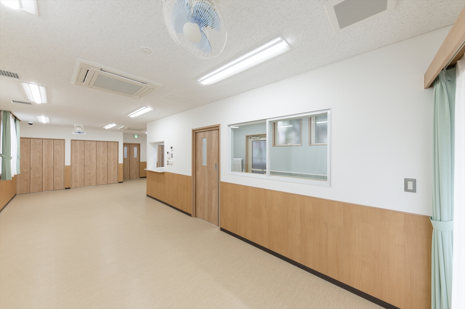 院内保育園の室内写真。木目調の壁と扉が並ぶ広いスペース、窓から光が入り明るく清潔な雰囲気の保育室。
