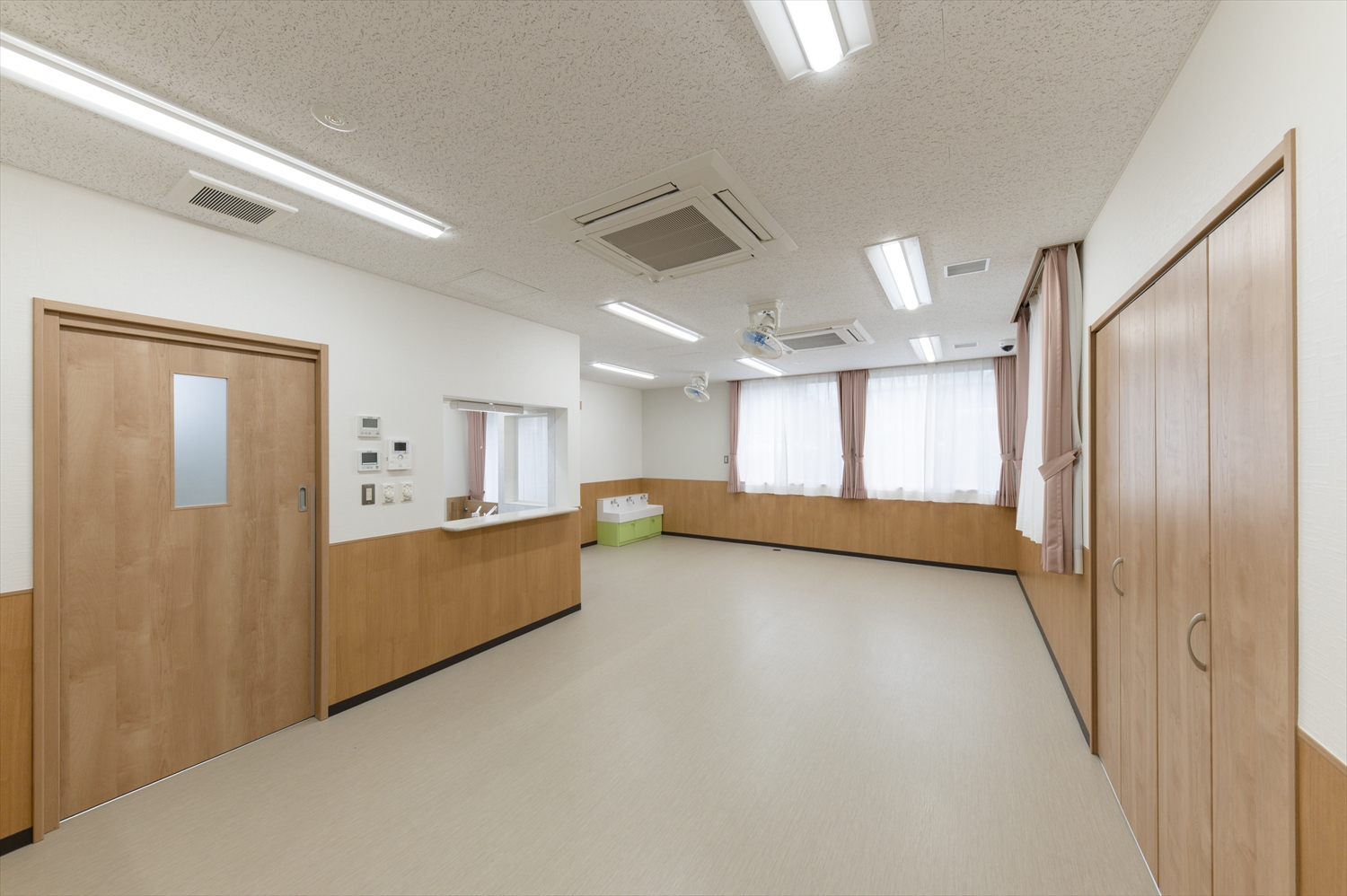 院内保育園の保育室。木目調の壁と扉で統一され、明るい窓から光が入る清潔で安心感のある室内。