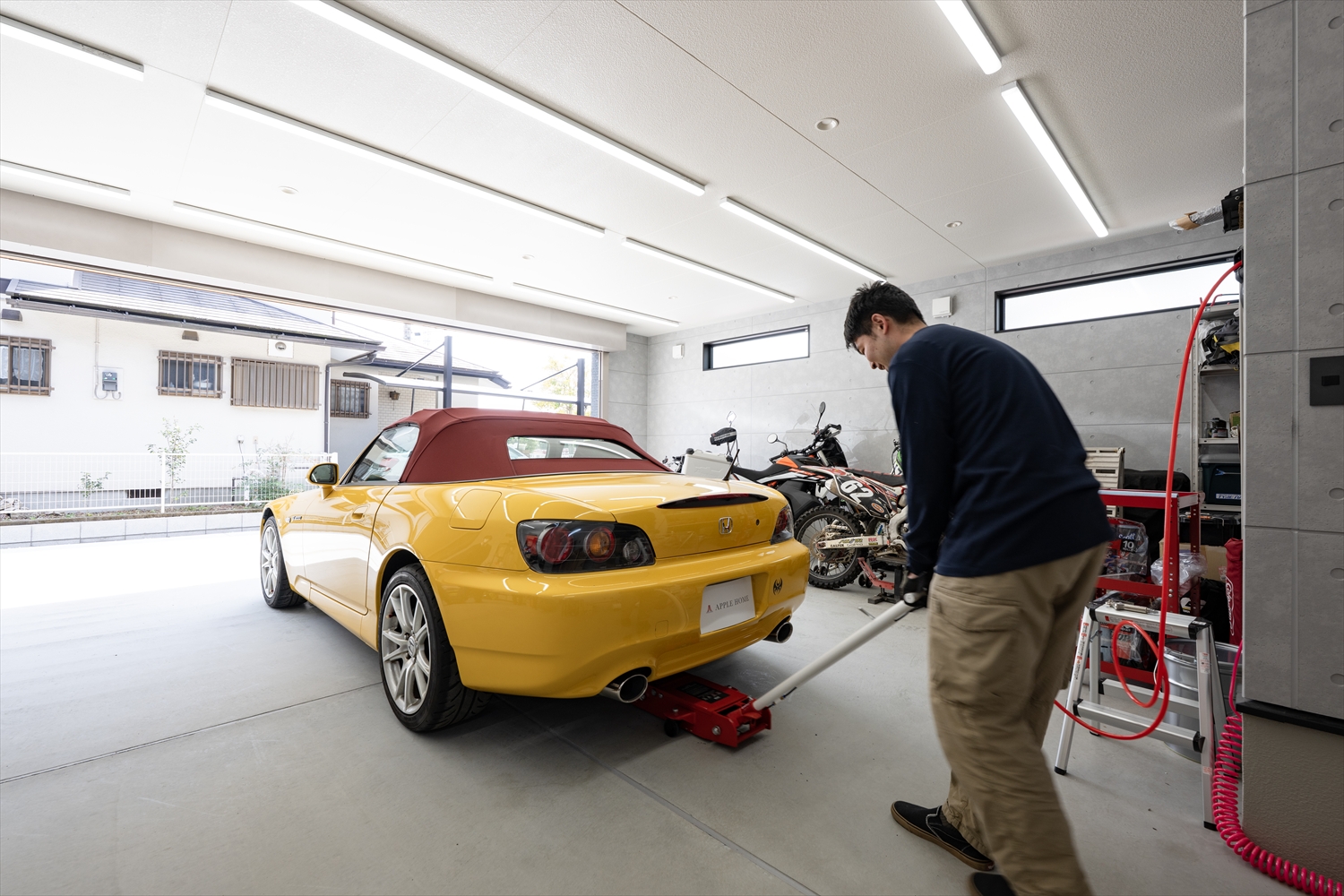 ビルトインガレージの大開口シャッター前で、車をジャッキアップして整備する様子(入間市のガレージハウス)