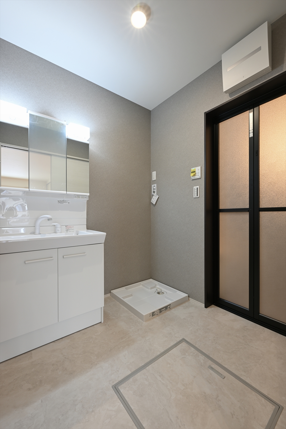 グレー系クロスの洗面脱衣室、三面鏡洗面台と洗濯機パンがある空間