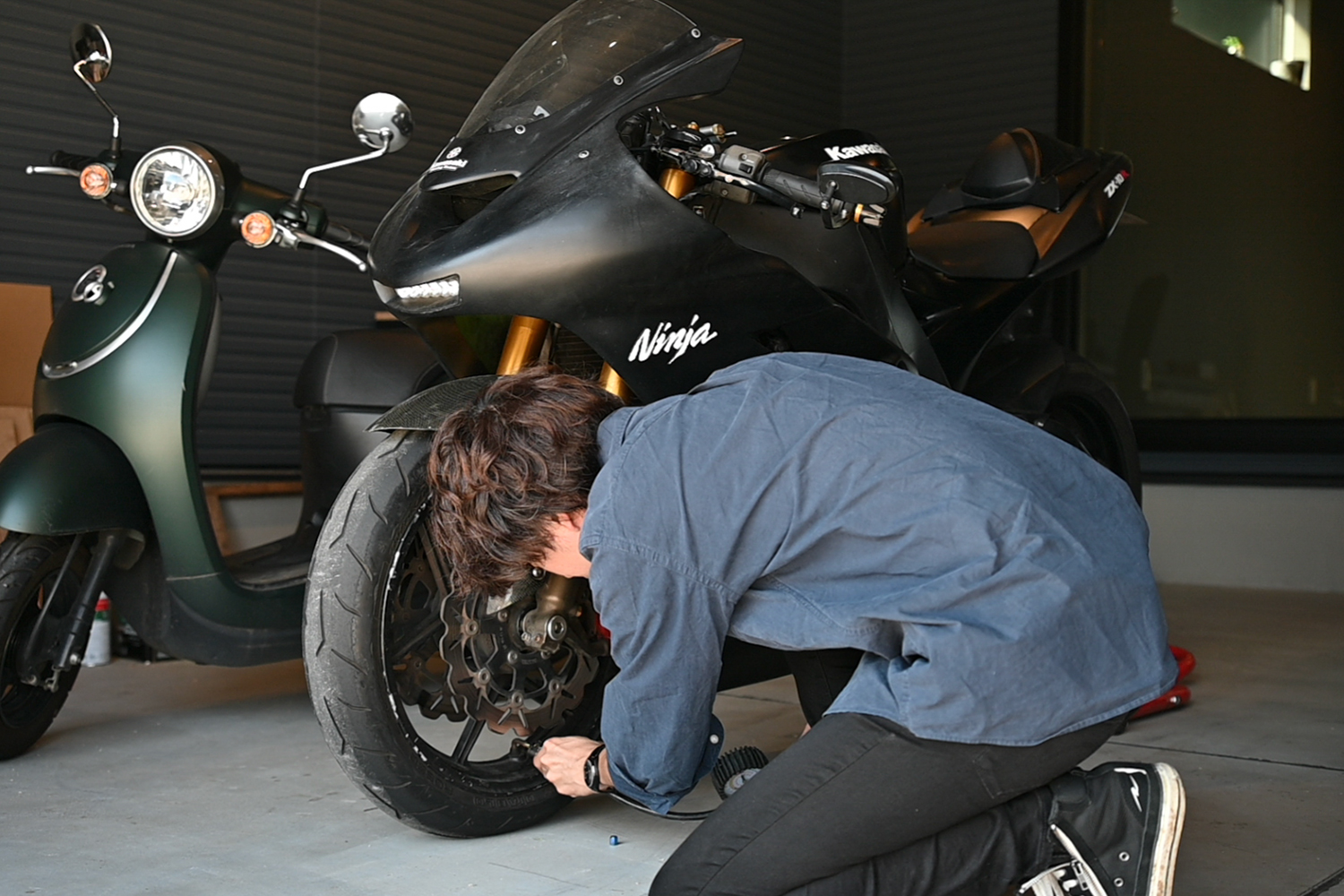 屋内のガレージでバイクの前輪付近に手をかけて作業する様子